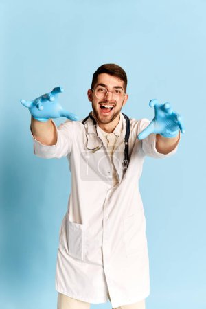 Photo for Funny doctor. Portrait of young cheerful man, doctor in white medical gown and protective gloves posing against blue studio background. Concept of medicine, profession, emotions, occupation. Ad - Royalty Free Image