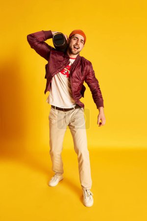 Photo for Full-length image of positive, handsome, young man in stylish casual clothes posing with music player against yellow studio background. Concept of emotions, male fashion, lifestyle, facial expression - Royalty Free Image