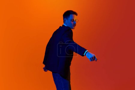 Photo for Looks delightful. Portrait of young cheerful man, student in jacket posing, dancing against orange studio background in neon light. Concept of youth, emotions, facial expression, lifestyle. Ad - Royalty Free Image