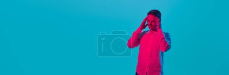 Photo for Young man suffering from stress and headache, holding head with hands, posing against blue studio background in pink neon light. Youth, emotions, facial expression concept. Banner. Copy space for as - Royalty Free Image