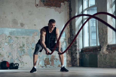 Photo for Strength, endurance and power. Muscular sportive young man doing functional exercises with battle ropes indoors. Fitness trainer. Concept of sportive lifestyle, body care, fitness, hobby, health - Royalty Free Image