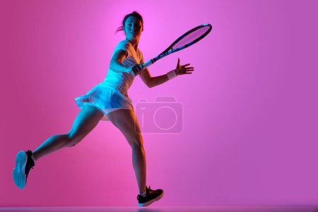 Téléchargez les photos : Image latérale pleine longueur de joueuse de tennis en mouvement, portant l'uniforme, s'entraînant sur fond de studio rose sous la lumière du néon. Concept de sport professionnel, mouvement, santé, action. Publicité - en image libre de droit