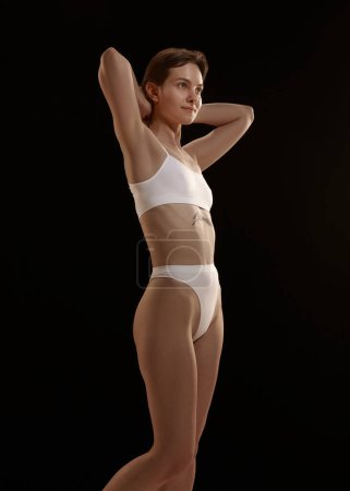 Photo for Slim, fit, young girl posing in white underwear against black studio background. Female model with tattoo on body. Concept of body and skin care, figure, fitness, health, wellness. - Royalty Free Image