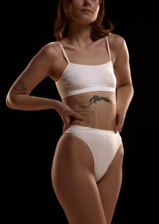 Photo for Cropped image of young woman with slim body in white underwear against black studio background. Tattoo, body art. Femininity. Concept of body and skin care, figure, fitness, health, wellness. - Royalty Free Image