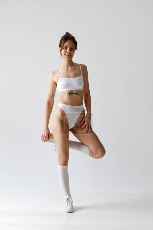 Photo for Full-length portrait of positive, young girl with slim, fit body posing in underwear, high socks and sneakers against grey studio background. Concept of body and skin care, figure, fitness, wellness. - Royalty Free Image
