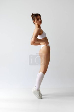 Photo for Full-length portrait of young girl with slim, fit body in underwear, high socks and sneakers against grey studio background. On tiptoe. Concept of body and skin care, figure, fitness, health, wellness - Royalty Free Image