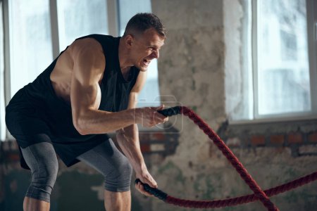 Photo for Coach. Young man with sportive, strong, muscular body keeping body toned, training with sports equipment, pulling battle ropes indoors. Concept of sportive lifestyle, body care, fitness, hobby, health - Royalty Free Image