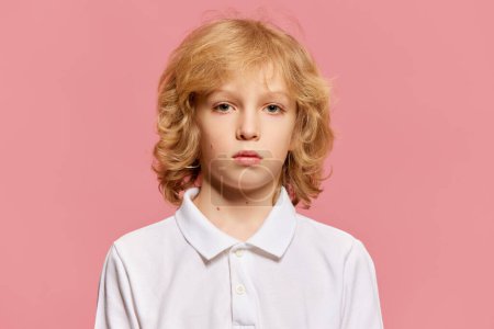 Photo for Portrait of little boy, child with curly blonde hair in white shirt attentively looking at camera against pink studio background. Concept of childhood, emotions, facial expression. Ad - Royalty Free Image