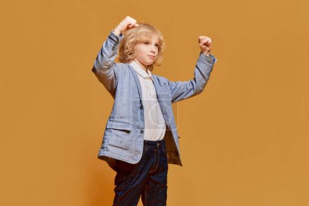 Photo for Winner. Success. Portrait of little boy, child with blonde curly hair posing in formal wear against orange studio background. Concept of childhood, emotions, facial expression, lifestyle. Ad - Royalty Free Image