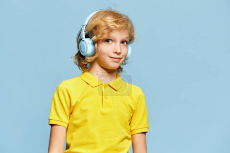 Téléchargez les photos : Petit garçon, enfant en chemise jaune écoutant de la musique dans des écouteurs, posant sur fond bleu studio. Concept d'enfance, émotions, expression faciale, mode de vie. Publicité - en image libre de droit