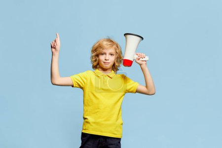 Photo for Little boy, child with blonde curly hair in yellow t-shirt rising finger, posing with megaphone against blue studio background. Concept of childhood, emotions, facial expression, lifestyle. Ad - Royalty Free Image