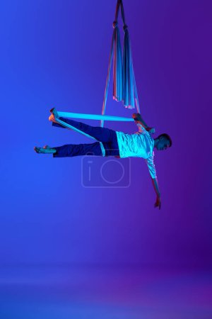 Téléchargez les photos : Homme jeune, fort et athlétique, entraînement professionnel de gymnaste aérien avec rubans de soie aériens sur fond violet bleu dégradé en lumière néon. Concept d'art, style de vie sportif, passe-temps, action et - en image libre de droit