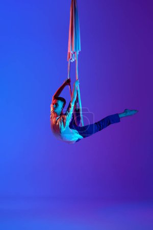 Foto de Actuación artística. Joven, acrobat entrenamiento con cintas aéreas, haciendo trucos de gimnasia contra el gradiente azul púrpura fondo en luz de neón. Concepto de arte, estilo de vida deportivo, acción, movimiento - Imagen libre de derechos