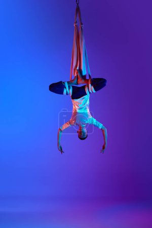 Photo for Young man, professional aerial acrobat, gymnast training with aerial ribbons against gradient blue purple background in neon light. Hanging upside down. Concept of art, sportive lifestyle and motion - Royalty Free Image