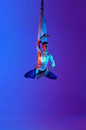 Foto de Hombre acróbata, profesional, gimnasta aérea artística colgando boca abajo en la seda aérea contra el gradiente azul púrpura fondo en luz de neón. Concepto de arte, estilo de vida deportivo, hobby, acción, movimiento - Imagen libre de derechos