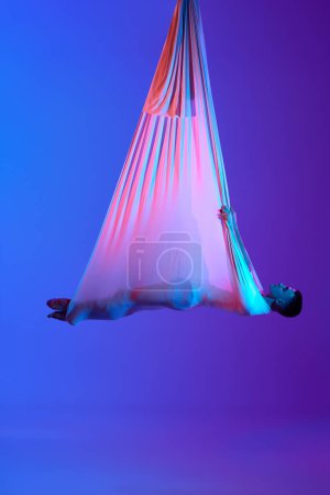 Foto de Hombre deportista, entrenamiento de gimnasta aérea profesional con hamaca, colgando en el aire contra el gradiente azul púrpura estudio de fondo en luz de neón. Concepto de arte, estilo de vida deportivo, acción y movimiento - Imagen libre de derechos