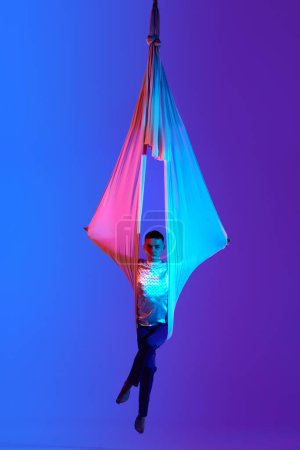 Foto de Hombre joven atlético, gimnasta aérea profesional haciendo trucos con cintas contra el fondo azul púrpura degradado en luz de neón. Mariposa. Concepto de arte, estilo de vida deportivo, hobby, acción y movimiento - Imagen libre de derechos