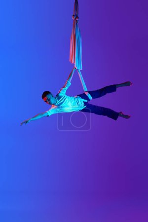 Foto de Acrobat, gimnasta aérea masculina profesional que cuelga boca abajo sobre seda aérea contra el fondo azul púrpura degradado en luz de neón. Concepto de arte, estilo de vida deportivo, hobby, acción y movimiento, belleza - Imagen libre de derechos