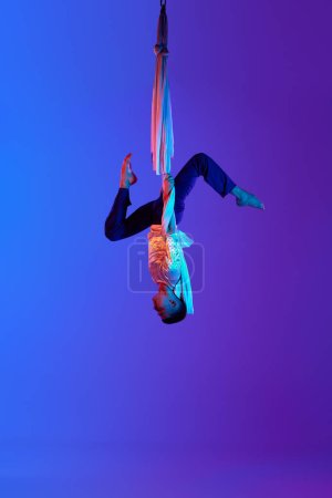 Foto de Gimnasta aérea masculina profesional colgando boca abajo en tela aérea contra el fondo azul púrpura degradado en luz de neón. Estética acrobática. Concepto de arte, estilo de vida deportivo, acción, movimiento - Imagen libre de derechos