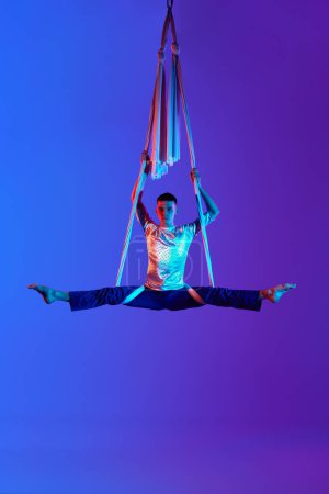 Foto de Entrenamiento profesional de gimnasta aérea masculina con cintas aéreas contra el fondo azul púrpura degradado en luz de neón. Posición del torzal y levitación. Concepto de arte, estilo de vida deportivo, acción, movimiento - Imagen libre de derechos