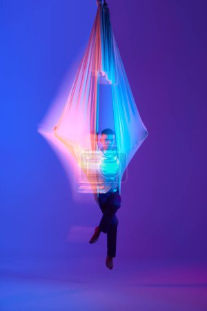 Foto de Hombre joven, acróbata aéreo, gimnasta de entrenamiento con tela aérea contra el fondo azul púrpura degradado en neón con luces mixtas. Concepto de arte, estilo de vida deportivo, hobby, acción y movimiento, belleza - Imagen libre de derechos