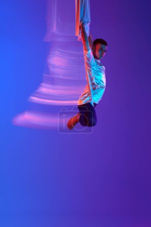 Foto de Hombre joven, gimnasta aérea, entrenamiento acróbata con tejido aéreo contra el fondo azul púrpura degradado en neón con luces mixtas. Concepto de arte, estilo de vida deportivo, hobby, acción y movimiento, belleza - Imagen libre de derechos