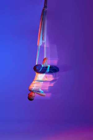 Foto de Joven, gimnasta aérea profesional haciendo trucos acrobáticos con cinta aérea contra el fondo azul púrpura degradado en neón con luces mixtas. Concepto de arte, estilo de vida deportivo, movimiento, belleza - Imagen libre de derechos