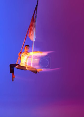 Foto de Hombre fuerte, gimnasta aérea profesional, entrenamiento acróbata con tejidos aéreos contra el gradiente azul púrpura fondo del estudio en neón con luces mixtas. Concepto de arte, estilo de vida deportivo, movimiento - Imagen libre de derechos