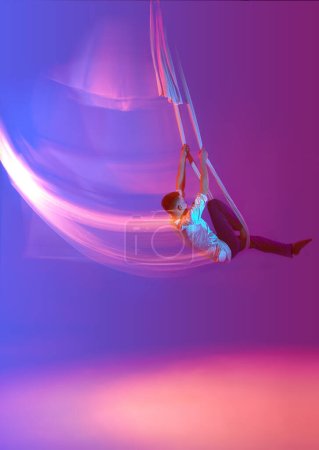 Téléchargez les photos : Jeune homme, acrobate d'entraînement avec des rubans aériens contre dégradé bleu violet fond studio au néon avec des lumières mélangées. Concept d'art, mode de vie sportif, passe-temps, action et mouvement, beauté - en image libre de droit