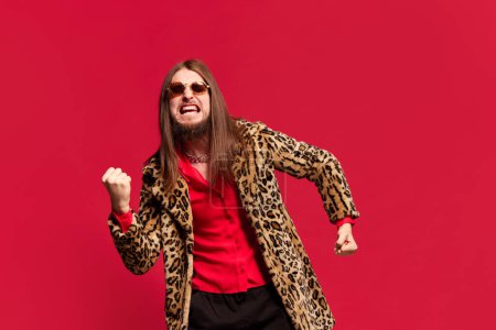 Photo for Portrait of young man with long hair posing in stylish fur coat and sunglasses against red studio background. Emotionally shouting. Concept of emotions, facial expression, lifestyle - Royalty Free Image