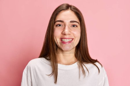 Téléchargez les photos : Portrait de jeune fille heureuse, souriante, en t-shirt blanc décontracté posant sur fond de studio rose. Je me sens bien. Ambiance positive. Concept de jeunesse, émotions, expression faciale, mode de vie - en image libre de droit