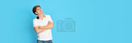 Foto de Sueños y fantasía. Joven hombre guapo en camiseta blanca posando con una cara pensativa sonriente sobre fondo azul del estudio. Concepto de emociones, juventud, expresión facial, estilo de vida. Copiar espacio para anuncio - Imagen libre de derechos