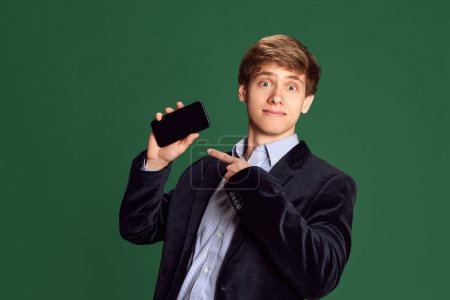 Photo for Portrait of young man in stylish jacket pointing on mobile phone screen against green studio background. Freelance. Concept of business, education, fashion, emotions, youth, lifestyle - Royalty Free Image