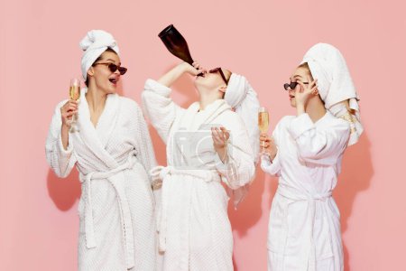 Foto de Reunión de spa. Tres hermosas chicas en albornoces bebiendo champán, divirtiéndose contra el fondo rosa del estudio. Celebración y disfrute. Concepto de juventud, belleza, amistad, fiesta, relajación - Imagen libre de derechos