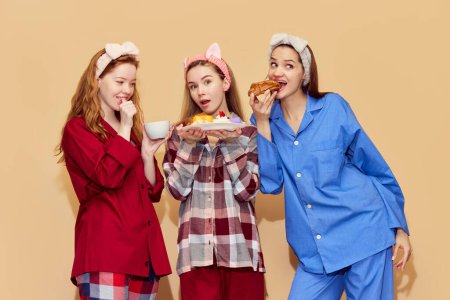 Téléchargez les photos : Une soirée pyjama. Jolies jeunes filles en pyjama confortable coloré mangeant des bonbons, s'amusant sur fond de studio orange. Concept de jeunesse, émotions, beauté, amitié, fête, détente - en image libre de droit