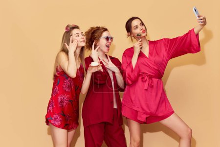 Foto de Hermosas chicas jóvenes en pijama rojo y túnicas tomando selfie con teléfono móvil contra el fondo del estudio. Diversión de pijamas. Concepto de juventud, emociones, belleza, amistad, fiesta, relajación - Imagen libre de derechos