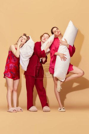 Photo for Sleepover with friends. Pretty young girls in pajamas holding pillows and yawning against studio background. Concept of youth, emotions, beauty, friendship, party, relaxation, fun, rest - Royalty Free Image
