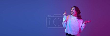 Photo for Excited, happy, positive young girl pointing with finger up against gradient blue purple studio background in neon light. Information. Concept of emotions, youth, lifestyle. Banner. Copy space for ad - Royalty Free Image