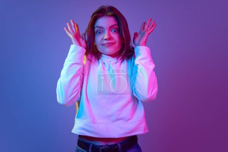Photo for Emotional young girl with big eyes looking at camera with questioning face, posing against gradient blue purple studio background in neon light. Emotions, youth, lifestyle, facial expression concept - Royalty Free Image