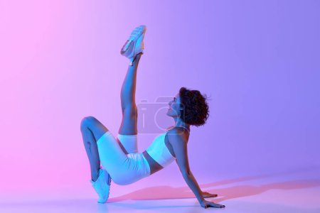 Foto de Mujer joven atlética en un cómodo entrenamiento de ropa deportiva, haciendo ejercicios de estiramiento contra el degradado fondo azul rosado en luz de neón. Estilo de vida deportivo, belleza, cuidado del cuerpo, fitness, concepto de salud - Imagen libre de derechos