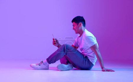 Photo for Happy, smiling young man in casual clothes sitting on floor, looking on mobile phone, texting messages against purple background in neon light. Concept of human emotions, youth, fashion, lifestyle - Royalty Free Image