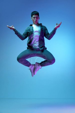 Foto de Relajación, calma. Joven hombre emotivo en ropa casual saltando en postura de yoga sobre fondo azul en luz de neón. Concepto de emociones humanas, juventud, moda, estilo de vida, sentimientos - Imagen libre de derechos