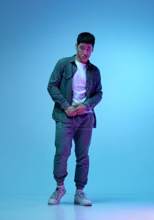 Photo for Full-length image of young asian guy in stylish casual clothes posing, looking with doubt and curiosity against blue background in neon light. Concept of human emotions, youth, fashion, lifestyle - Royalty Free Image