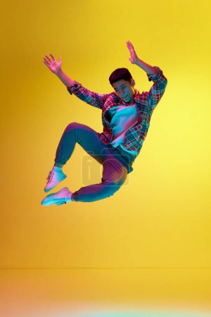 Photo for Delightful, reaching success. Full-length image of young man in casual clothes jumping against yellow background in neon light. Concept of human emotions, youth, fashion, lifestyle - Royalty Free Image