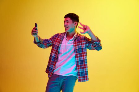 Téléchargez les photos : Positif, souriant, jeune homme prenant selfie avec téléphone portable, posant sur fond jaune sous la lumière du néon. Influence des médias sociaux. Concept d'émotions humaines, de jeunesse, de mode, de style de vie - en image libre de droit