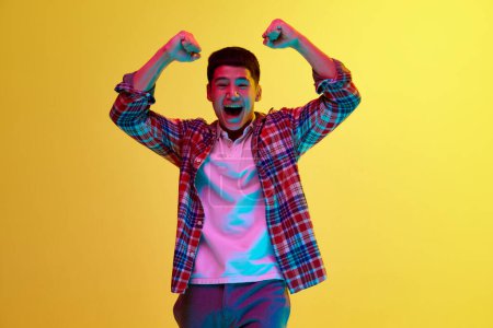 Photo for Young korean guy in stylish casual clothes showing emotions of success, win against yellow background in neon light. Achievement. Concept of human emotions, youth, fashion, lifestyle - Royalty Free Image