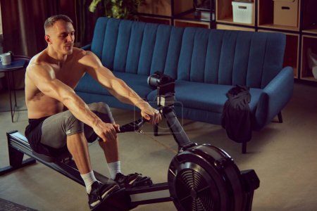 Photo for Strong, sportive, shirtless man with muscular body doing exercises with stationary rowing machine. Home gym. Keeping body in tone. Concept of sportive lifestyle, body and health care, fitness, health - Royalty Free Image