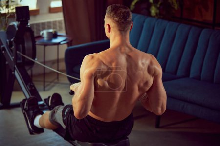 Photo for Rear view of young sportive man with relief, strong, muscular back, body training at home, doing exercises with stationary rowing machine. Concept of sportive lifestyle, body and health care, fitness - Royalty Free Image