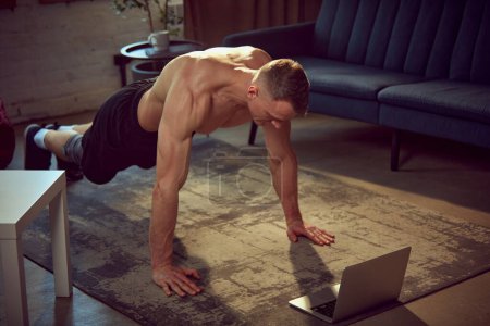 Photo for Young sportive man with strong, muscular body training shirtless, doing push-up exercises at home. Online training with laptop. Concept of sportive lifestyle, body and health care, fitness, health - Royalty Free Image