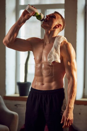 Photo for Handsome muscular man with relief strong body, standing shirtless, drinking water after training session at home on daytime. Concept of sportive lifestyle, body and health care, fitness, health - Royalty Free Image